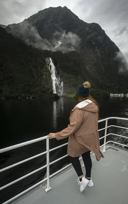 tasha amy in a brown jacket and pom-pom beanie stands on a boat deck, admiring bowen falls plunging from a mountain into the fiord at Milford Sound.
