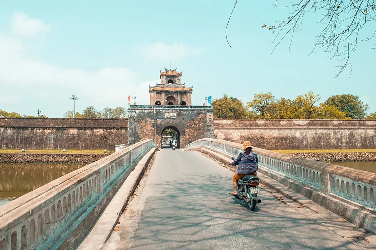 3 Weeks In Vietnam Itinerary: The Perfect First-Time Route 68 Motorcyclist crossing a bridge leading to a historic gate in Hue, Vietnam