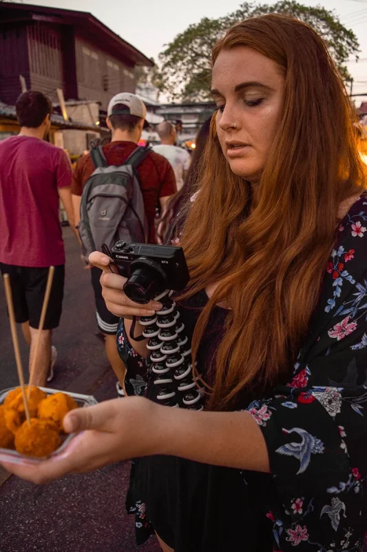 Tasha Amy at the Sunday Chiang Mai Market focused on her camera while holding a plate of Thai fried balls, with crowds in the background