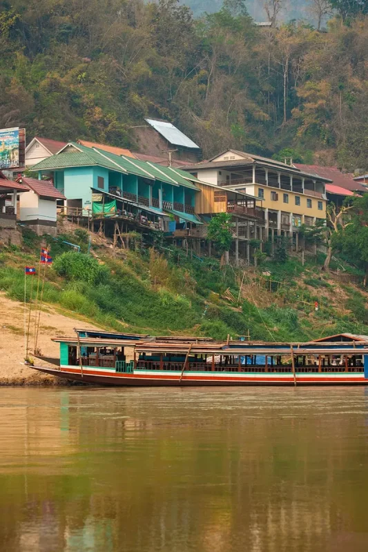 Slow Boat Laos: My Honest Guide After Doing It Twice (2026) 48 A slow boat sits parked up below colorful houses on the Mekong River