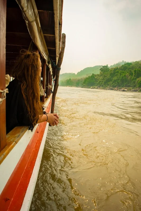 Slow Boat Laos: My Honest Guide After Doing It Twice (2026) 28 Tasha Amy rests her hands on the edge of a slow boat to Laos as she looks out over the Mekong River