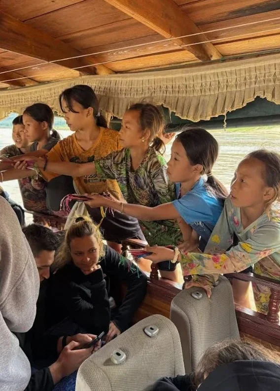 Slow Boat Laos: My Honest Guide After Doing It Twice (2026) 40 Local children reaching inside the slow boat while its parked up at a village trying to sell bracelets to tourists in Laos