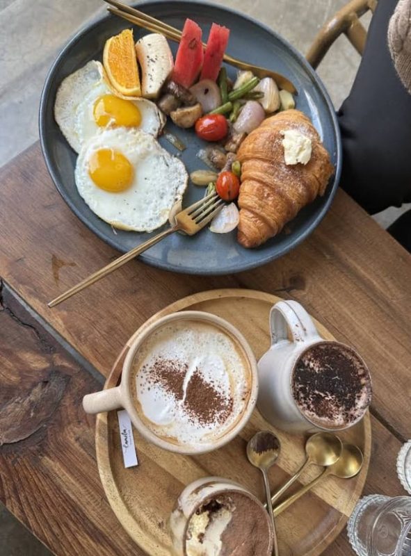 Best Restaurants In Pokhara: Cafes, Cheap Eats & Local Food 24 fried eggs, roasted vegetables and a crossiant on one plate with a hot chocolate and coffee on a wooden board at Cafe Camellia in Pokhara