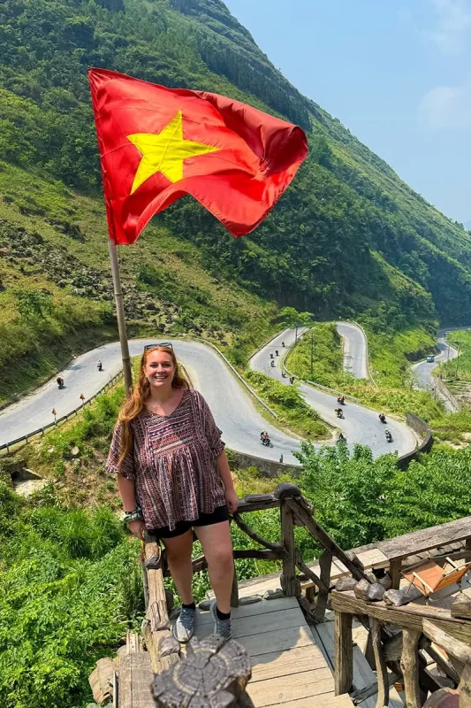 Tasha beside a Vietnamese flag overlooking the winding road at Tham Ma Pass