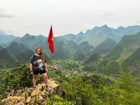 Tasha standing beside a Vietnamese flag at a mountain viewpoint on my Ha Giang Loop itinerary