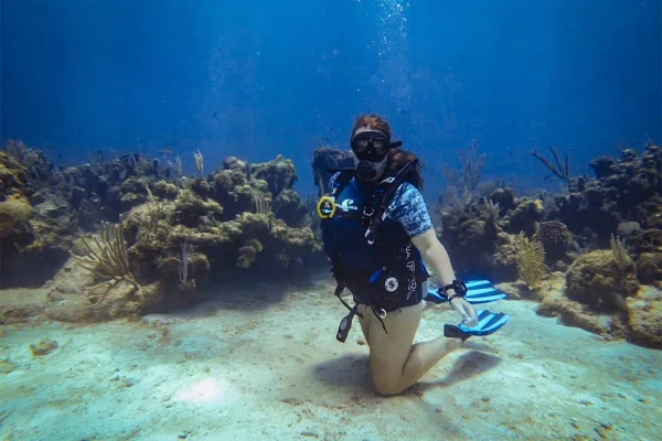 Tasha Amy with blue fins kneeling on a sandy patch of seabed surrounded by reef after learning to dive in Roatan
