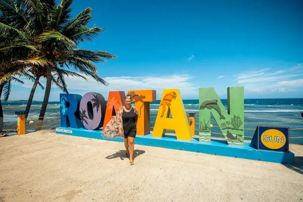 Tasha Amy standing in front of the colorful Roatan sign beside palm trees and the ocean