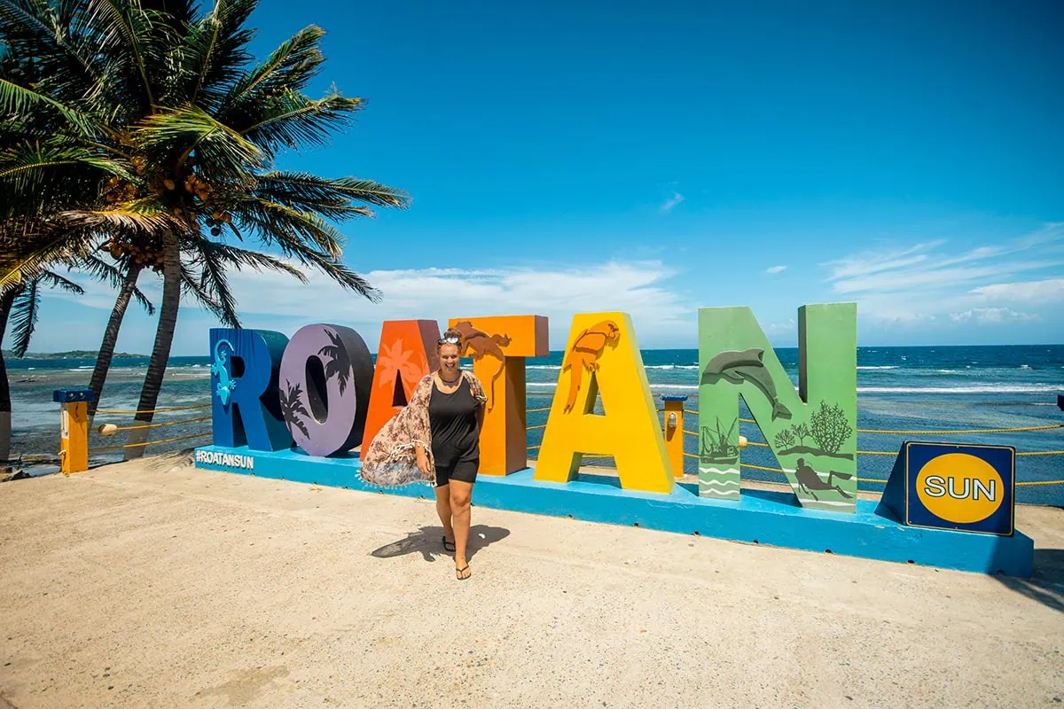 Tasha Amy standing in front of the colorful Roatan sign beside palm trees and the ocean