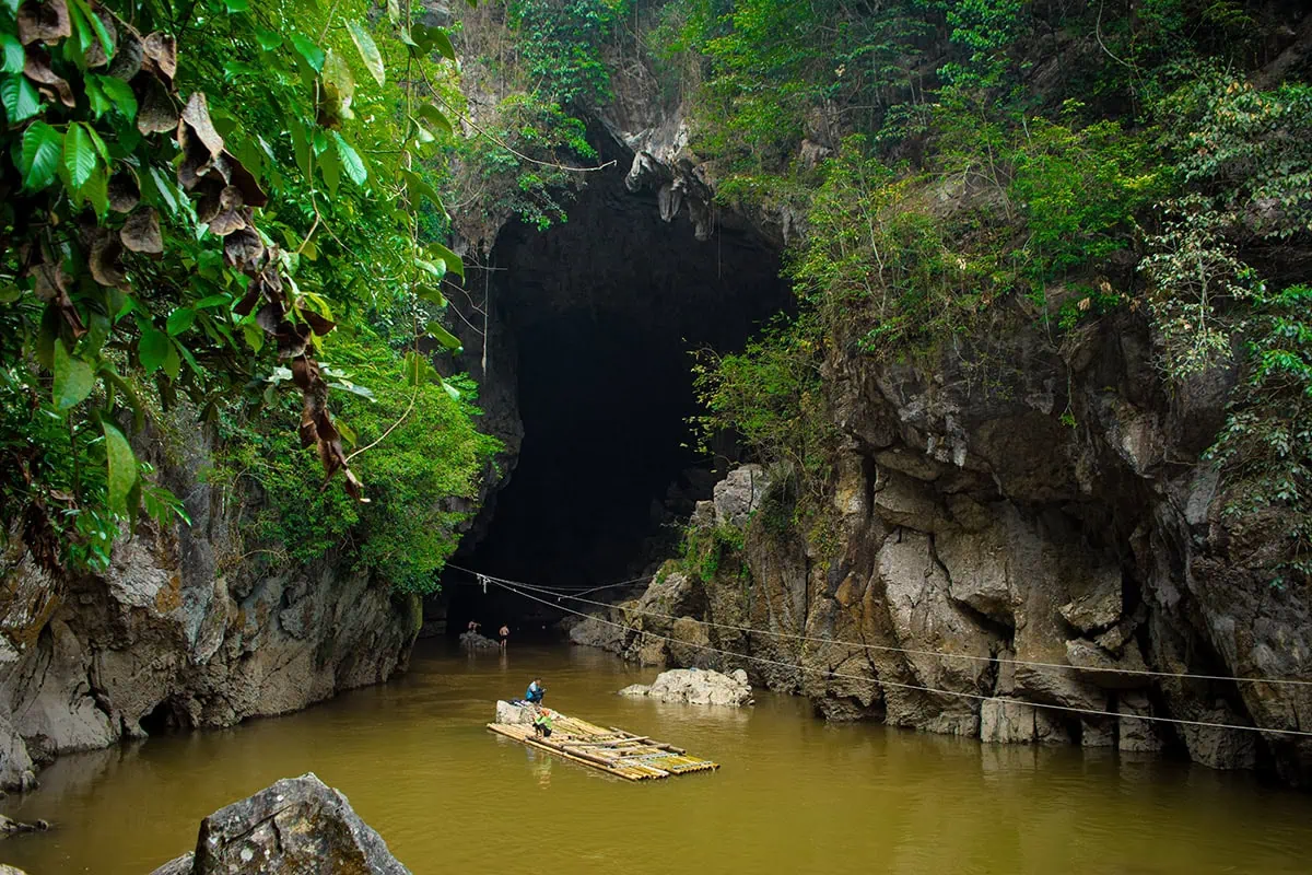 Bamboo raft approaching a dark Hang Tham Lin cave entrance surrounded by rocky cliffs and forest