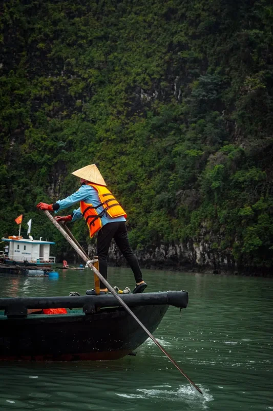 Luon Cave Ha Long Bay: Is It Worth Visiting? 62 Rower wearing an orange life jacket and conical hat guiding a wooden boat beside limestone cliffs near Luon Cave