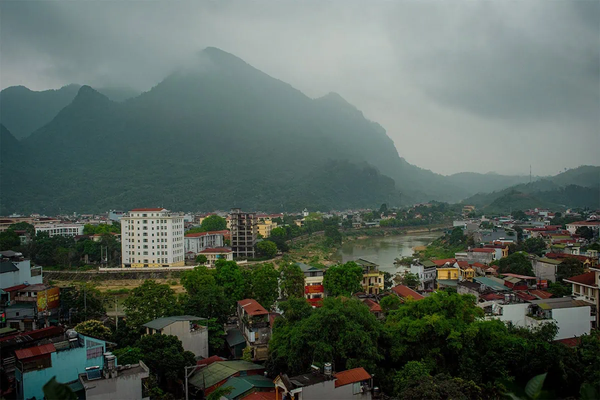 Best Hostels In Ha Giang: Where To Stay Before The Loop 56 Buildings beside the river in Ha Giang City, with green mountains rising into low clouds from Top of the Loop Hostel