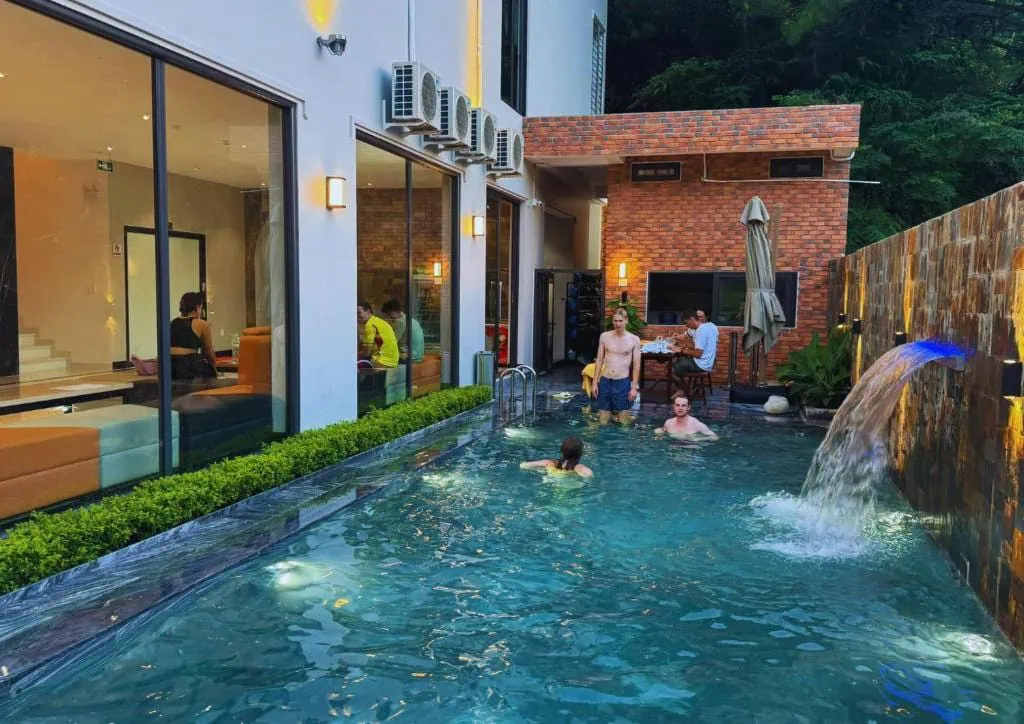 Best Hostels In Ha Giang: Where To Stay Before The Loop 63 Travelers swimming in an outdoor pool at Ha Giang Hostel, with a wall-mounted waterfall beside the lounge