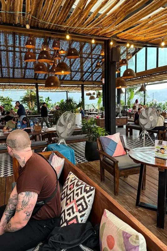 Best Hostels In Ha Giang: Where To Stay Before The Loop 71 Rooftop restaurant at Top Of The Loop Hostel with wooden seating, woven pendant lights, plants and travelers