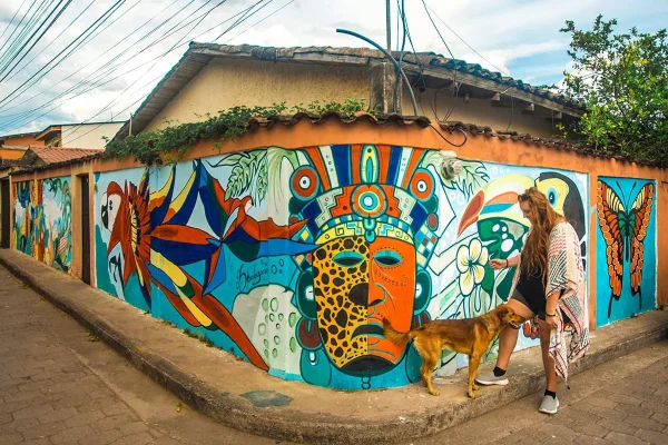 Tasha Amy and dog beside a colorful mural featuring Maya-inspired designs, tropical birds, and a butterfly in Copan Ruinas