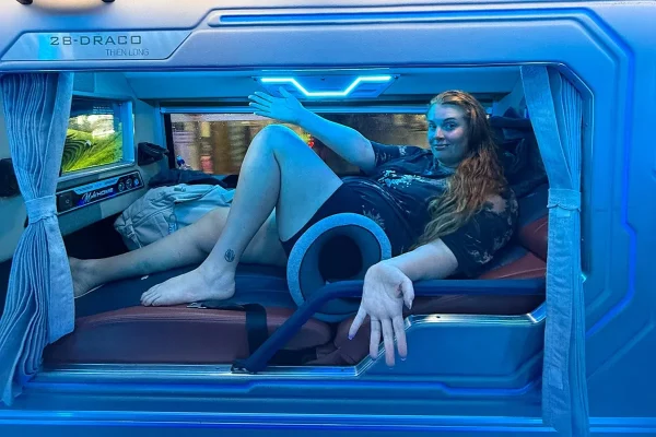 Tasha Amy reclining in an individual sleeper bus cabin with blue lighting and open privacy curtains