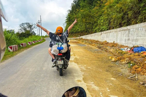 Tasha Amy wearing an orange helmet and raising both arms while riding with an Easy Rider on the Ha Giang Loop