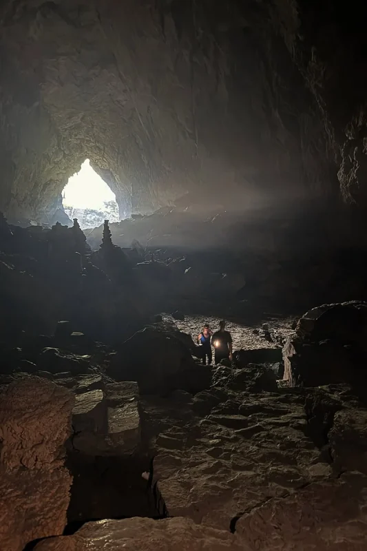 Two travelers using phone lights on a rocky path inside the enormous Ta Lung Cave chamber