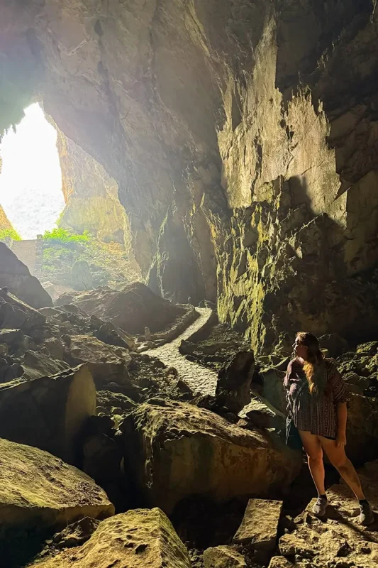 Tasha Amy standing among large rocks near the sunlit entrance of Ta Lung Cave