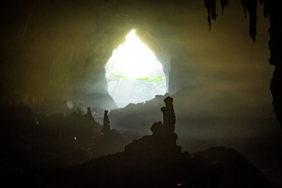 Daylight streaming through a cave opening behind silhouetted stalagmites inside Ta Lung Cave